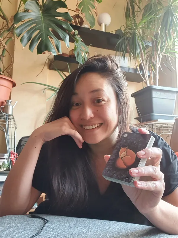 Isabelle smiling, cheek resting on one hand, holding a deck showing Fox, with plants behind her.