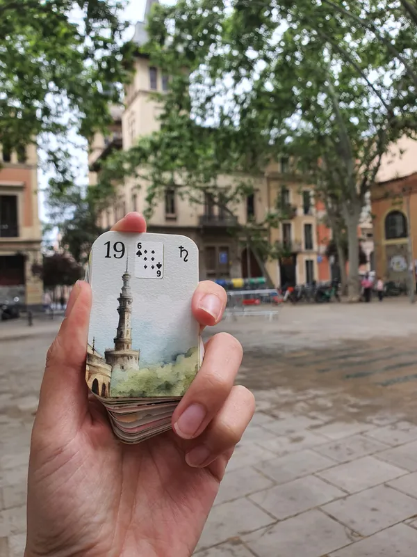 Tower, card 19 from the Aquarelle deck, held in front of a tree-lined square and a building with a tower.